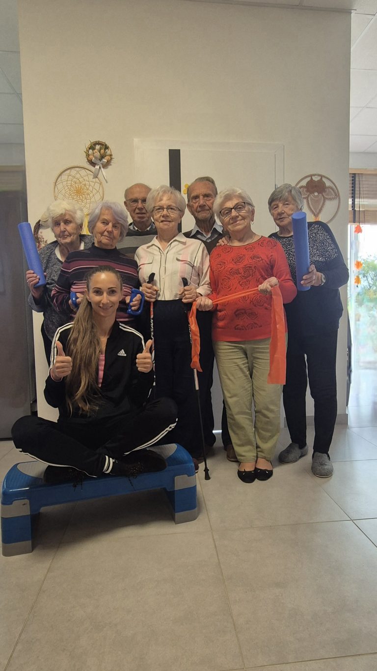 Grupa starszych osób oraz jedna młoda instruktorka pozuje do zdjęcia w pomieszczeniu. Młoda kobieta w czarnym stroju sportowym siedzi na niebieskim stopniu do aerobiku i unosi kciuki. Starsze osoby (trzy kobiety i trzech mężczyzn) stoją za nią, trzymając niebieskie rolki piankowe, pomarańczową taśmę elastyczną lub kijki do nordic walkingu.